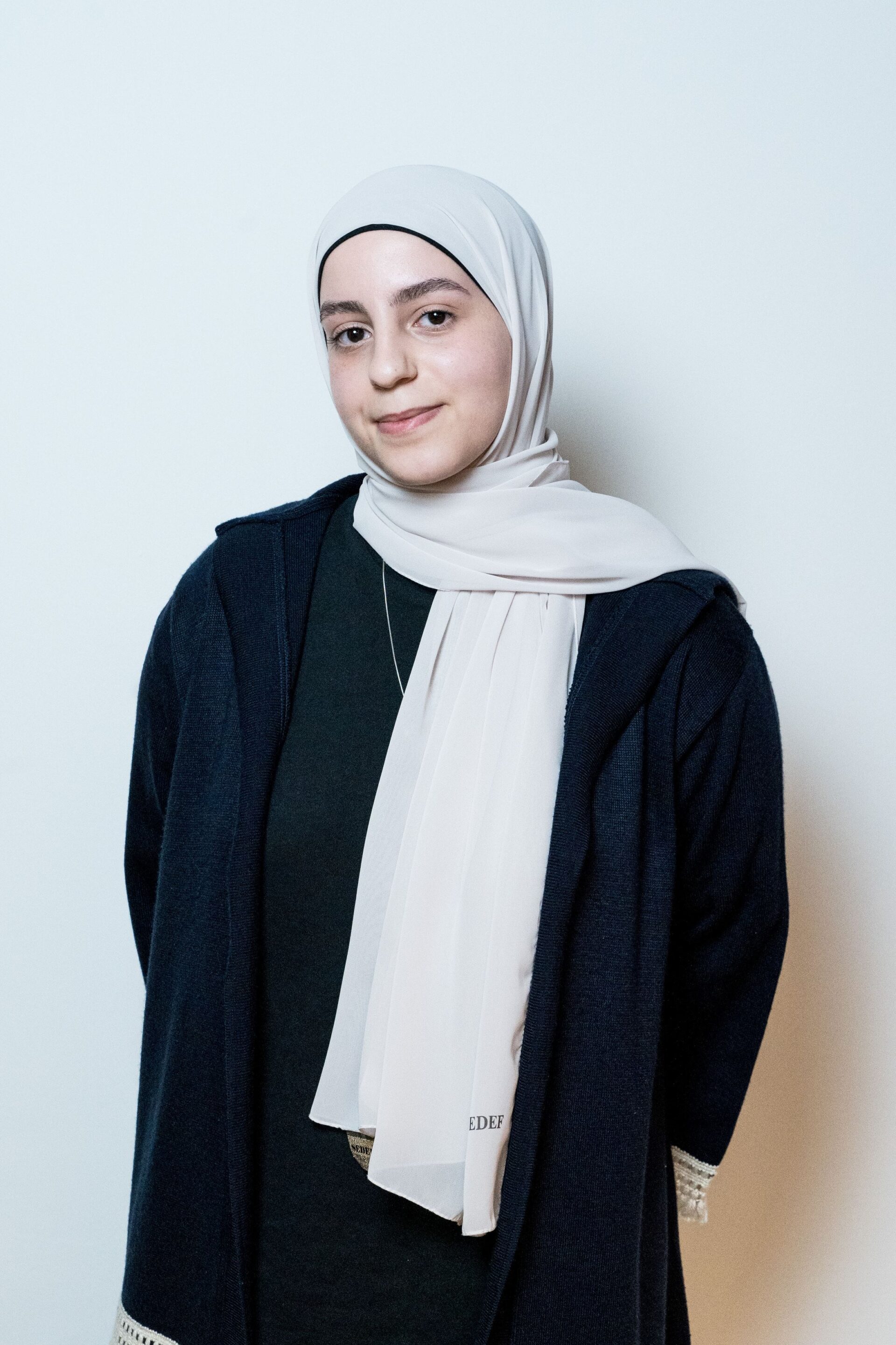 A woman wearing a light gray hijab and a dark coat stands against a pale background.