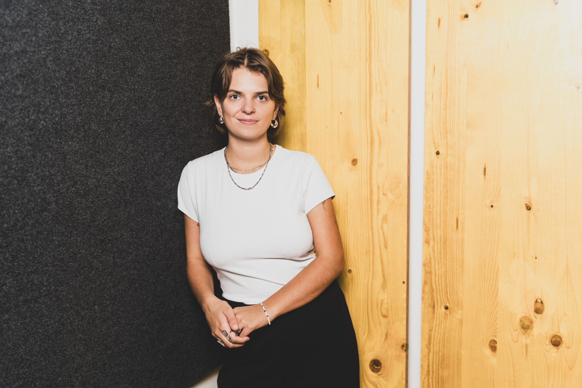 Portrait of a woman with short hair wearing a white t-shirt and a black skirt, standing against a dark wall and wooden panels.